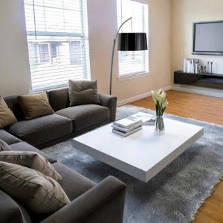 A modern living room featuring a gray sofa, white coffee table, and wall-mounted TV, with large windows providing natural light.