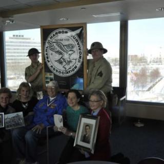 A group of people posing indoors with historical photographs and costumes, accompanied by a large emblem displayed in the background.