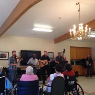 A band performs for an audience in a cozy indoor setting, with several attendees in wheelchairs.