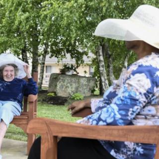 Two people sitting on benches in a park, wearing large hats and engaging in conversation.