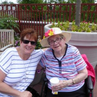 Two women sitting outside, smiling, with one in a wheelchair wearing a floral hat.