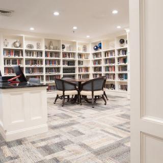 A cozy library with white bookshelves, a central table surrounded by chairs, and bright overhead lighting.