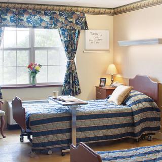 A shared room in a nursing home featuring two beds with patterned bedspreads, a window with floral curtains, and a bedside table with flowers.