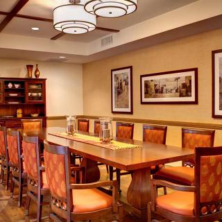A modern dining room with a long wooden table and orange cushioned chairs, featuring elegant lighting and framed wall art.