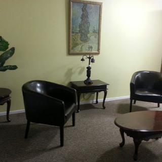 A waiting room with black leather chairs, a wooden coffee table, and a decorative lamp on a side table beneath a painting.