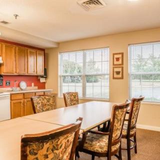 A cozy dining area with a wooden table and patterned chairs, adjacent to a kitchen with warm wooden cabinetry and large windows.