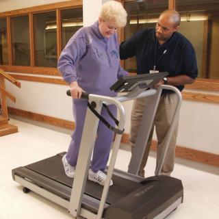 An elderly woman receiving assistance while walking on a treadmill for physical therapy.