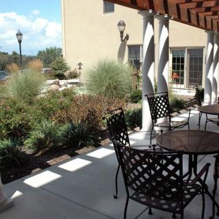 An outdoor patio area with tables and chairs beneath a pergola, surrounded by landscaped greenery on a sunny day.