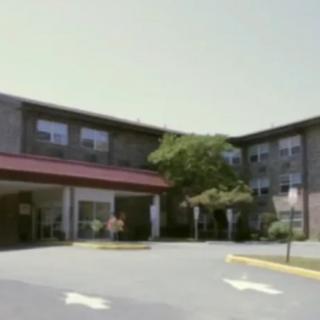 A three-story building with a red awning and surrounding trees, adjacent to a parking lot.