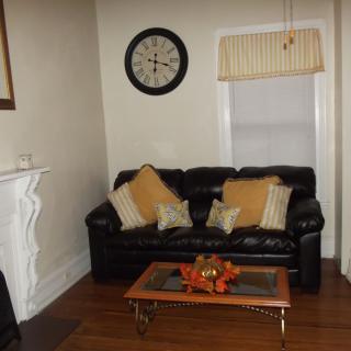 A cozy living room with a black leather sofa, decorated with pillows, a wooden coffee table, an elegant clock on the wall, and a fireplace.