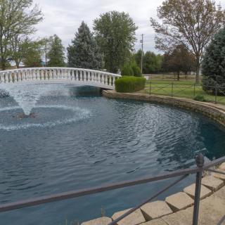 A serene park scene featuring a pond with a fountain and a decorative bridge surrounded by greenery.