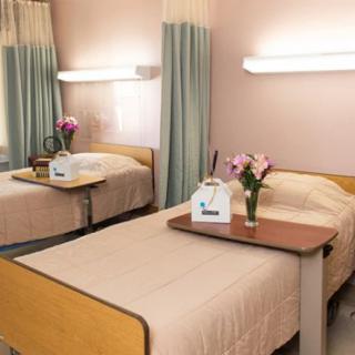 A hospital room with two neatly made twin beds, equipped with tray tables and adorned with flower arrangements.