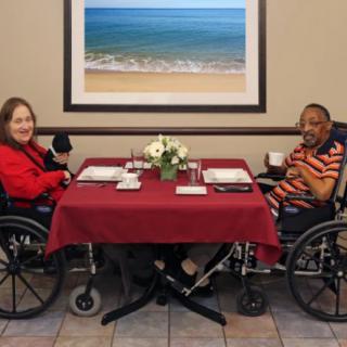 Two people sitting at a dining table in wheelchairs, enjoying coffee beneath a framed beach scene.