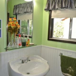 A cozy bathroom with green walls, featuring a sink, mirror, and window adorned with flowers and decor.