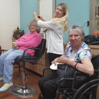 A hairdresser styling the hair of an elderly woman while another woman in a wheelchair smiles at the camera.