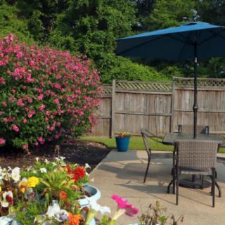 A cozy backyard patio with an umbrella, table, and chairs surrounded by vibrant flowers and greenery.