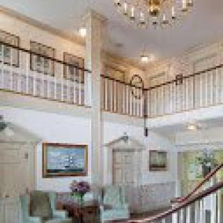 A traditional interior featuring a grand staircase, elegant chandelier, and cozy seating area with artwork adorning the walls.