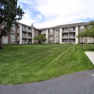 A three-story apartment building with a well-maintained lawn and trees under a partly cloudy sky.