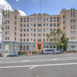 A large urban building with a street view, featuring several vehicles parked in front and trees lining the sidewalk under a partly cloudy sky.