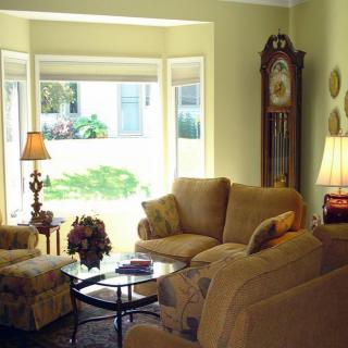 A cozy living room with comfortable seating, a grandfather clock, and ample sunlight streaming through a bay window.