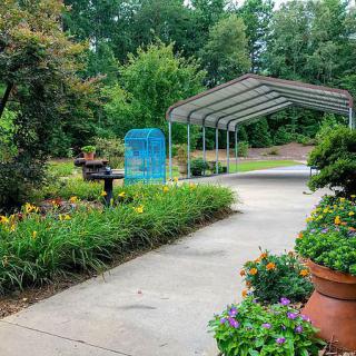 A scenic garden pathway lined with lush greenery and potted plants, leading to an outdoor structure surrounded by trees.