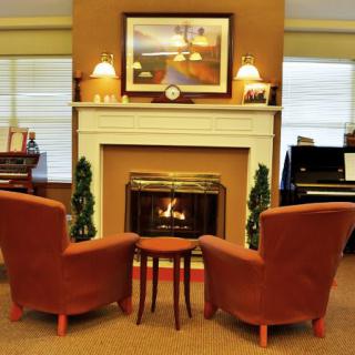 A cozy living room with two chairs facing a lit fireplace, flanked by lamps and decorated with artwork.