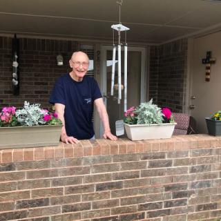 A man smiling while standing on a porch with flowers and a wind chime, leaning on a brick wall.