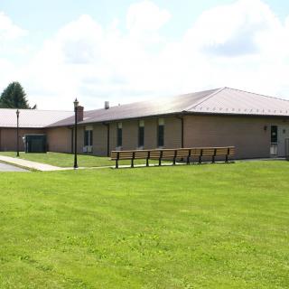 A single-story brick building with a metal roof, surrounded by a well-maintained lawn and benches in the foreground.