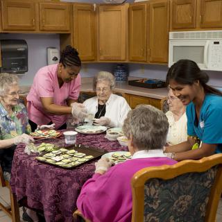 Caregivers assisting elderly women during a meal in a kitchen setting.