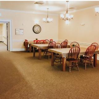 A spacious dining room with wooden tables and chairs, illuminated by chandeliers, featuring a carpeted floor and a wall-mounted clock.