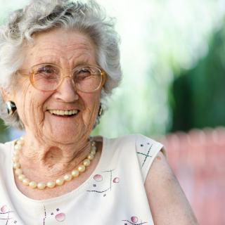 An elderly woman with a warm smile, wearing glasses and a pearl necklace, enjoys a sunny day outdoors.