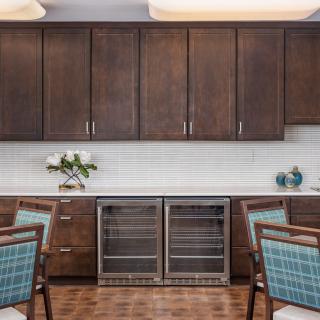 A modern kitchen with dark wooden cabinets, a sleek countertop, and dual wine fridges, accented by decorative flowers and chairs.