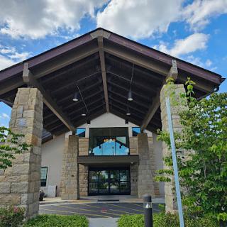 A modern architectural entrance featuring a large overhanging roof and stone columns, surrounded by greenery under a bright sky.