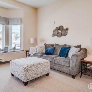 A cozy living room with a gray sofa, plush ottoman, and decorative cushions, bathed in natural light from a large window.