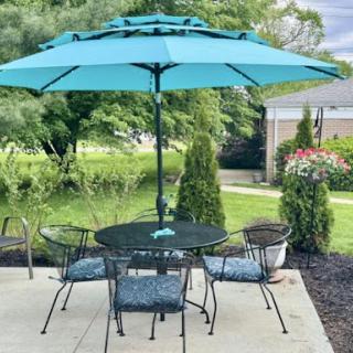 A tranquil outdoor patio setup featuring a teal umbrella over a table surrounded by chairs, set amid lush greenery.