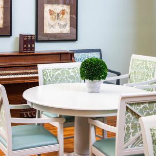 A cozy dining area with a round table and chairs, complemented by artwork and a piano in the background.