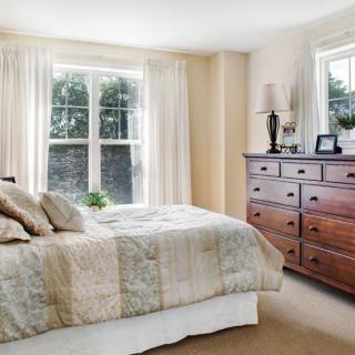 A cozy bedroom with a large bed and wooden dresser next to sunlit windows.
