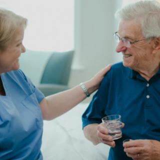 A nurse provides care and support to an elderly patient, who is holding a glass of water.
