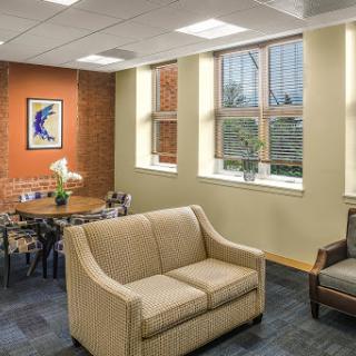 A cozy office lounge area featuring soft seating, a dining table, and large windows allowing natural light.