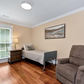 A cozy bedroom features a single bed, an armchair, and a nightstand with a lamp on a wooden floor.