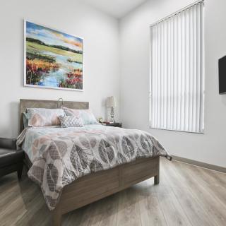 A modern bedroom featuring a bed with patterned bedding, a landscape painting above the headboard, and tall window blinds filtering light into the room.