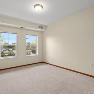A spacious, empty room with two windows allowing natural light to flood in, featuring a carpeted floor and neutral wall colors.