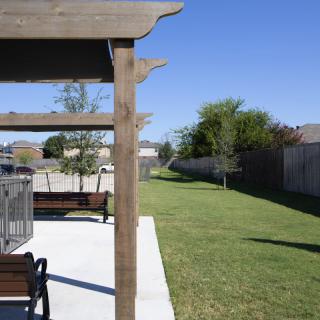 A sunny outdoor area featuring a pergola, benches, and a well-maintained lawn with trees.