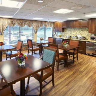 A cozy dining room with wooden tables and chairs, decorated with flowers and adjacent to a kitchen area.