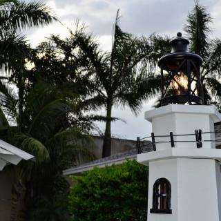 A small lighthouse with a glowing lamp stands against a backdrop of palm trees in the evening.