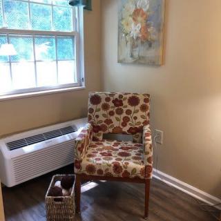A cozy corner featuring a floral-patterned chair next to a window, with a decorative painting on the wall and a basket on the floor.