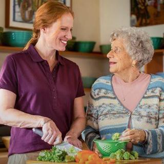 A younger woman and an elderly woman smile while preparing vegetables together in the kitchen.