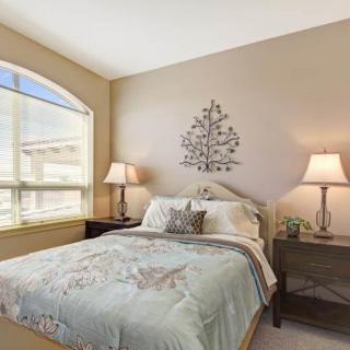 A cozy bedroom with natural light, featuring a bed with patterned bedding, two bedside lamps, and decorative wall art.