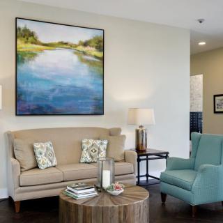 A cozy living room with a beige sofa, blue armchair, wooden coffee table, and landscape artwork on the wall.