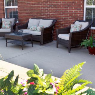 A cozy outdoor patio featuring wicker furniture, including two sofas and a chair, arranged around a coffee table with decorative plants nearby.